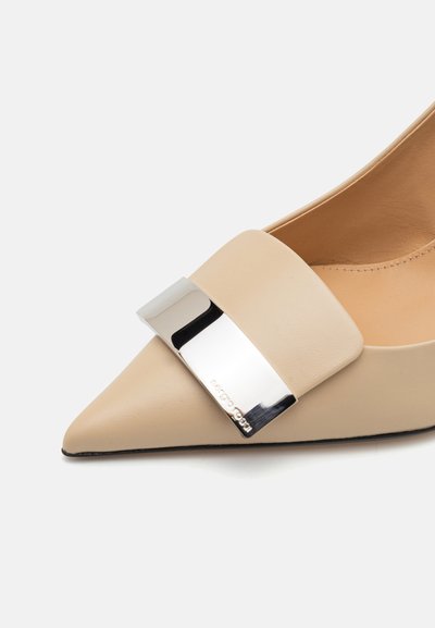 Beige läder sko med spetsig tå, bred rem och silverfärgad metallspänne som accent. Slät textur med svart gummisula.