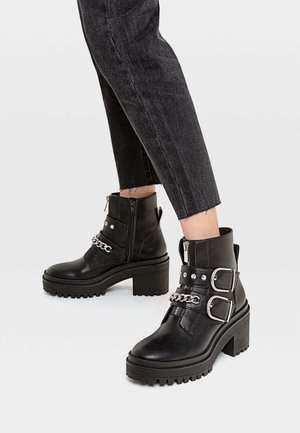 Sorte ankelstøvler med chunky hæle, sølvspænder og kædedetaljer, båret med korte sorte jeans.