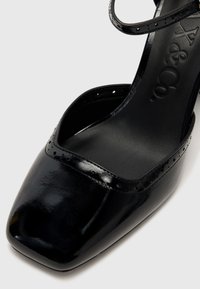 Schwarzer Lacklederschuh mit quadratischer Zehenform, dekorativen perforierten Akzenten entlang der Kante und einem schmalen Knöchelriemen.