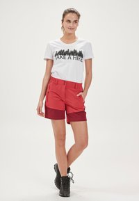 Röda vandringsshorts i lätt material, med en kontrasterande mörkare midja och fickor, i kombination med en vit t-shirt som har svart text.