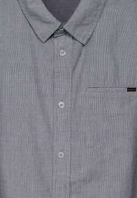 Grå knäppskjorta i strukturerad tyg, med krage, två knappar och en vänster bröstficka med en liten etikett.