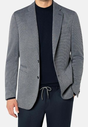 Mand iført en grå tekstureret blazer over en sort sweater og marineblå bukser med en grå snor, stående med den ene hånd i lommen.