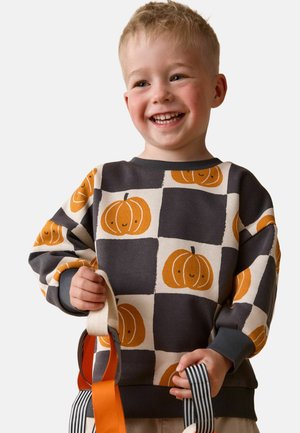 Smiling young child in a pumpkin-patterned sweater holding striped fabric straps, standing against a plain white background.
