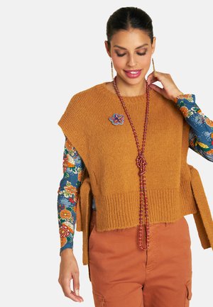Senfgelber Strickpullover mit kurzen Ärmeln und seitlichen Schlitzen, über einem gemusterten blumigen Langarmshirt getragen. Eine Perlenkette ergänzt den Look.