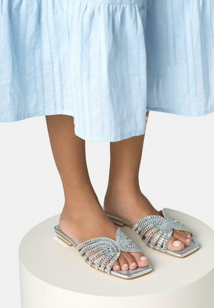 Pies descalzos con sandalias planas plateadas de tiras, decoradas con adornos cuadrados, de pie sobre una plataforma redonda blanca con una falda azul claro.