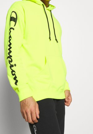 Personne portant un sweat à capuche Champion jaune néon avec un logo noir sur la manche, associé à un pantalon noir avec un petit logo près de la poche.