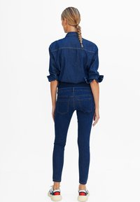 Camisa de mezclilla oscura con mangas enrolladas, combinada con jeans skinny de mezclilla oscura. Bolsillos traseros visibles y detalles en los puños; el modelo lleva zapatillas casuales.