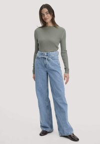 Hellgraues geripptes Longsleeve-Oberteil kombiniert mit hellblauen hochgeschnittenen weit geschnittenen Jeans, die ein Stoffband in der Taille und dunkle Schuhe haben.