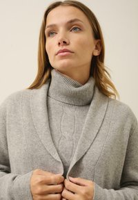 Cardigan de punto gris con cuello de chal, superpuesto a un cuello de tortuga acanalado. Presenta una textura suave y sutiles detalles de diseño.