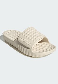 Adidasin liukukengät vaaleanbeigessä, joissa on tekstuuri päällä, joka näyttää kuplamaisilta kuvioilta, leveä hihna ja uritettu pohja, joka tarjoaa pitoa.