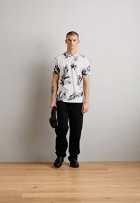 Witte t-shirt met zwart tropisch bladenpatroon, gecombineerd met zwarte broek en schoenen. Model houdt een zwarte tas vast en staat op een houten vloer.