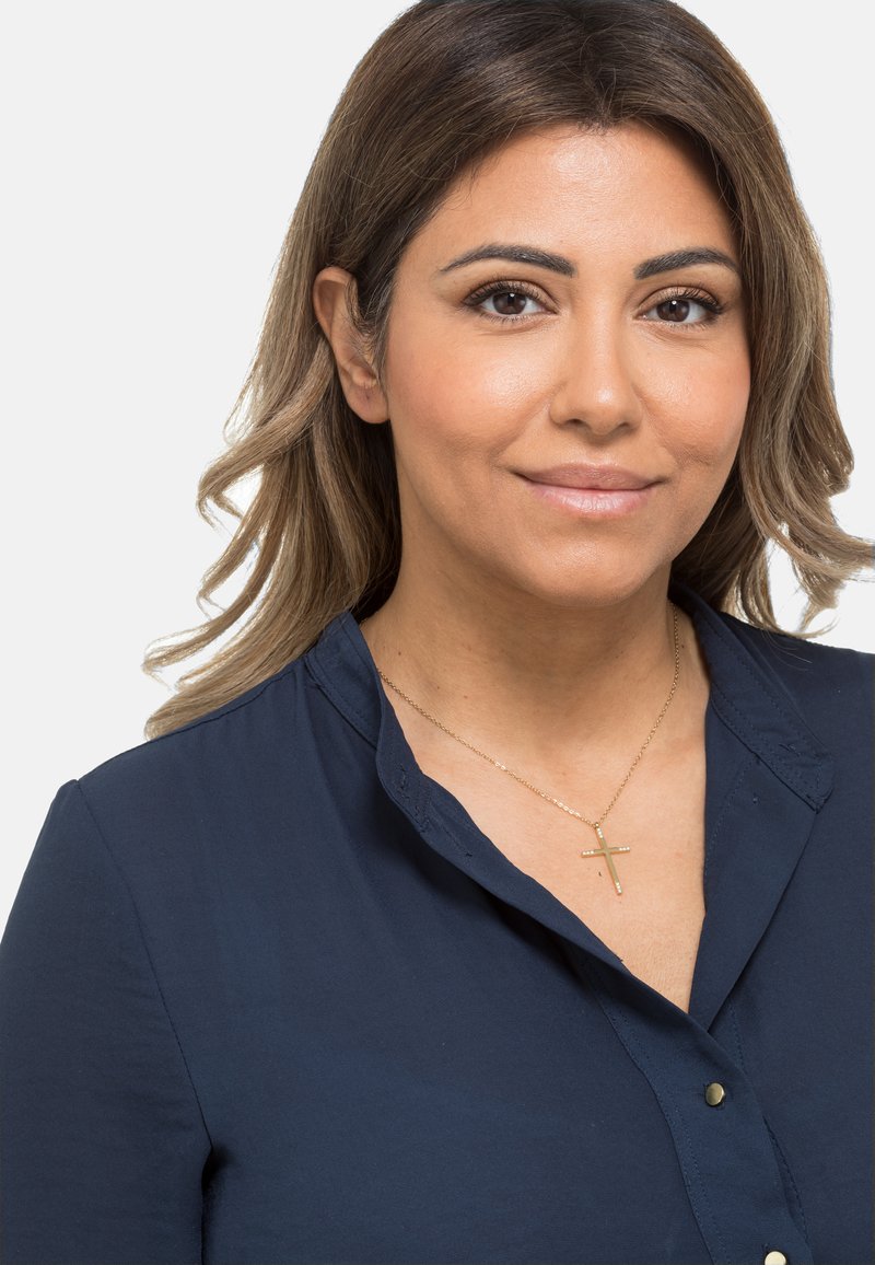 Marineblauwe overhemeld met kraag, gemaakt van soepele stof en voorzien van een gouden kruis ketting. Haar gestyled in losse golven. Neutrale achtergrond.