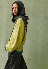 Gul strikket sweater med grå hætte, ribbede manchetter og en grøn lagdelt skjorte underneath, parret med mørkegrå løstsiddende jeans.