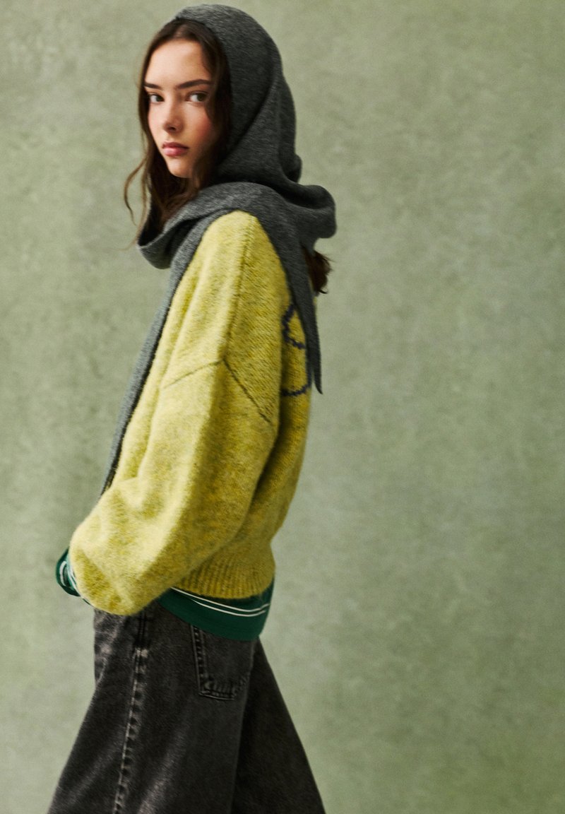 Gul strikket sweater med grå hætte, ribbede manchetter og en grøn lagdelt skjorte underneath, parret med mørkegrå løstsiddende jeans.