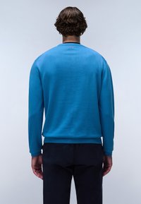 Blauwe gebreide trui met lange mouwen en geribbelde manchetten, voorzien van een klein logo-detail op de achterkant van de halslijn, gecombineerd met donkere broeken.