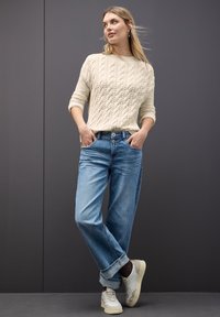 Cremfarbener Zopfstrickpullover, lässig sitzende hellblaue Jeans und weiße Sneakers. Das Outfit hat einen casual Stil mit umgeschlagenen Säumen an den Hosen.