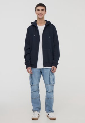 Granatowa bluza rozpinana z kapturem i sznurkiem, noszona na białej koszuli, w zestawie z jasnoniebieskimi spodniami cargo. Ubranie uzupełniają białe trampki.