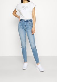 Camiseta blanca con la impresión "Apoya tu planeta local", combinada con pantalones vaqueros de cintura alta en azul claro y zapatillas blancas. Diseño casual y sencillo.
