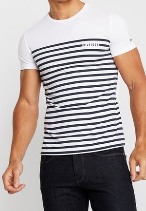 Mężczyzna noszący biało-granatowy t-shirt w paski z krótkim rękawem z napisem "HILFIGER" oraz ciemne jeansy, kadrowany na wysokości szyi i talii.