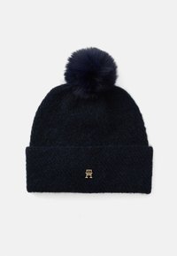 Marine gebreide beanie met een zachte pompon bovenop en een omgeslagen rand. Heeft een klein goudkleurig logo aan de voorkant. De textuur lijkt fuzzy.