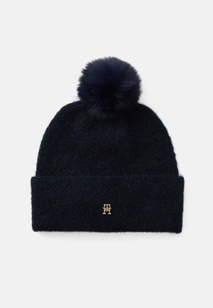 Navy knitted beanie with a soft pom-pom on top and a folded cuff. Features a small gold logo on the front. Texture appears fuzzy.