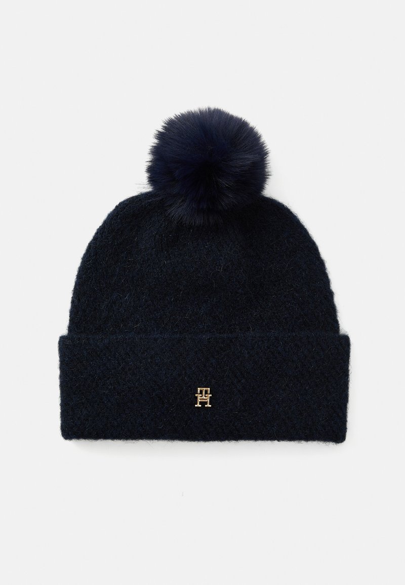 Marine gebreide beanie met een zachte pompon bovenop en een omgeslagen rand. Heeft een klein goudkleurig logo aan de voorkant. De textuur lijkt fuzzy.
