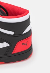 Wysokie sneakersy z czarną syntetyczną cholewką, czerwonymi akcentami, białymi paskami i teksturowanym kołnierzem. Posiadają czarne logo Pumy z boku.