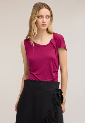 Mujer con cabello largo y rubio lleva una blusa de manga corta de color rosa oscuro y una falda negra cruzada con un lazo atado al lado, mirando hacia su izquierda.