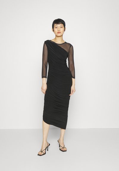 Czarna sukienka z marszczeniem, z długimi przezroczystymi rękawami, asymetrycznym dekoltem i dopasowanym krojem. Modelka nosi czarne sandały na obcasie z paskami.