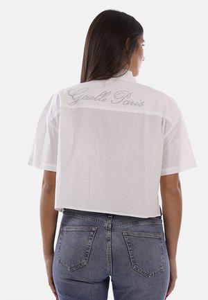 Donna con capelli lunghi e scuri che indossa una maglietta corta bianca a maniche corte con la scritta "Guelle Paris" sulla schiena e jeans di denim blu.