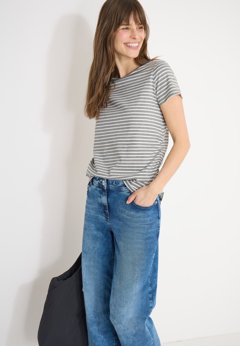 Femme souriante, portant un t-shirt rayé gris et blanc avec un jean bleu, debout contre un mur blanc et tenant un sac noir.