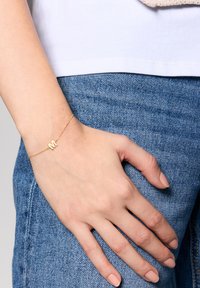 Złota bransoletka łańcuszkowa z dekoracyjnym charmem w kształcie litery "M", noszona na ręce spoczywającej na dżinsie, prezentująca gładką teksturę i prosty design.