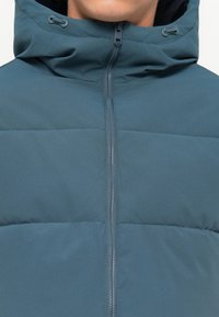 Padded jacket in deep teal with a textured finish, featuring a hood and central zipper. Stitched panels create a structured appearance.