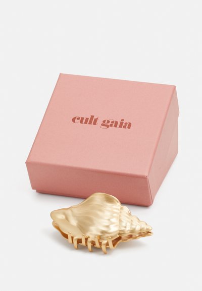Clip para el cabello en forma de concha de color dorado, colocado junto a una caja rosa con "cult gaia" impreso en marrón. Presenta bordes curvados y un diseño texturizado.
