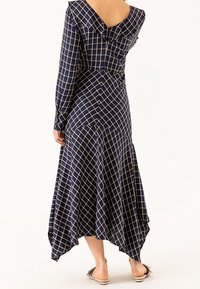 Kobieta w długiej, niebieskiej w kratę sukience z długim rękawem, o asymetrycznym dole i odkrytych plecach, w połączeniu z beżowymi klapkami.