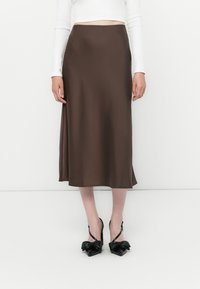 Smeđa satin midi suknja s glatkom teksturom i bočnim prorezom, uparena s crnim štiklama koje imaju naglaske u obliku mašne i strukturirani dizajn.