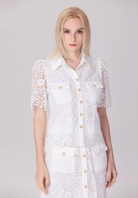 Robe chemise en dentelle blanche avec des manches courtes à volants, un col, des boutons à l'avant, et deux poches poitrine avec des accents beiges.