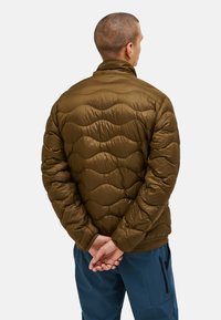 Giacca puffer verde oliva con un design trapuntato, colletto alto e finitura texturizzata. Indossata sopra pantaloni blu navy, vista da dietro.