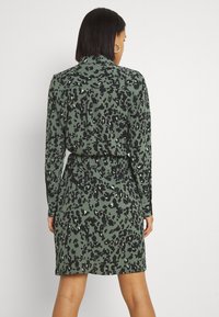 Grön leopardmönstrad klänning med långa ärmar, krage och insvängd midja. Tillverkad av lätt tyg med en slät textur.