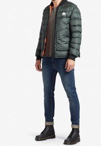 Zelená péřová bunda se oranžovým zipem a logem, černými manžetami; nošena přes hnědý triko, v kombinaci s modrými džínami a černými šněrovacími botami.