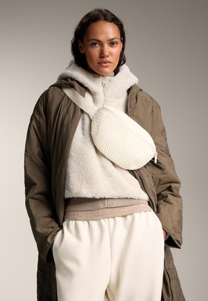 Witte gestructureerde crossbodytas gedragen over een beige fleece hoodie en een olijfgroene gewatteerde jas; gecombineerd met crème elastische broek met tailleband.