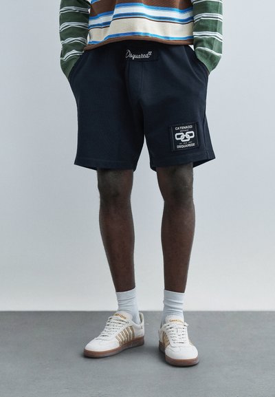 Personne portant un short noir Dsquared2 avec un écusson logo, une chemise à manches longues rayée, des chaussettes blanches et des baskets beiges, debout sur un sol gris.