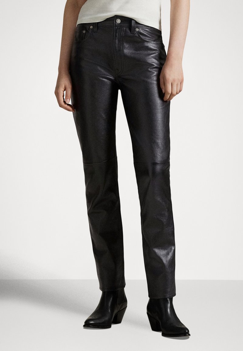 Άτομο που φοράει μαύρο δερμάτινο παντελόνι και μαύρα μποτάκια με τακούνι, στέκεται μπροστά σε απλό φωτεινό φόντο.