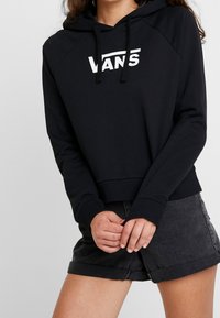 Donna che indossa una felpa nera Vans con lacci e pantaloni scuri in denim arrotolati, con le mani intrecciate in vita.