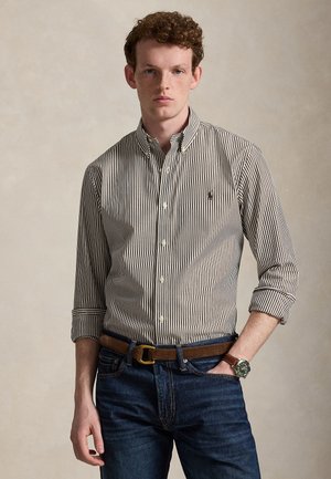 Ung mand med krøllet hår iført en brun og hvidstribet skjorte med knapper, mørke jeans, brun bælte og armbåndsur, stående med den ene hånd i lommen.