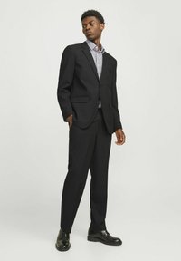 Schwarzer Anzug mit einem strukturierten Blazer und geraden Hosen, getragen über einem blau-weißen gestreiften Hemd, ergänzt durch schwarze Lederschuhe.