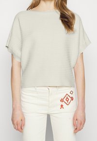 Light grey, short-sleeved sweater with ribbed texture, rounded neckline. Paired with cream-colored jeans featuring orange geometric embroidery.