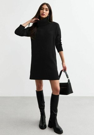 Robe noire en tricot avec un col montant, des manches longues et un ourlet côtelé. Associée à des bottes noires jusqu'aux genoux et un petit sac à main noir.