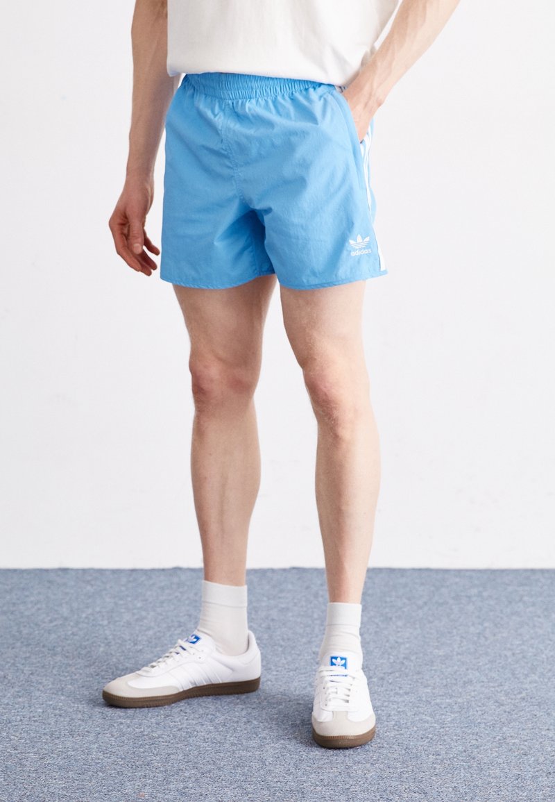 Světle modré sportovní šortky s elastickým pasem, zdobené bílými bočními pruhy a logem Adidas na levém lemu.