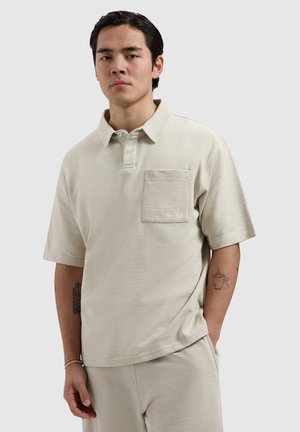 Jonge man met donker haar, gekleed in een beige korte-mouw polo en bijpassende broek, staand met één hand in zijn zak tegen een effen achtergrond.